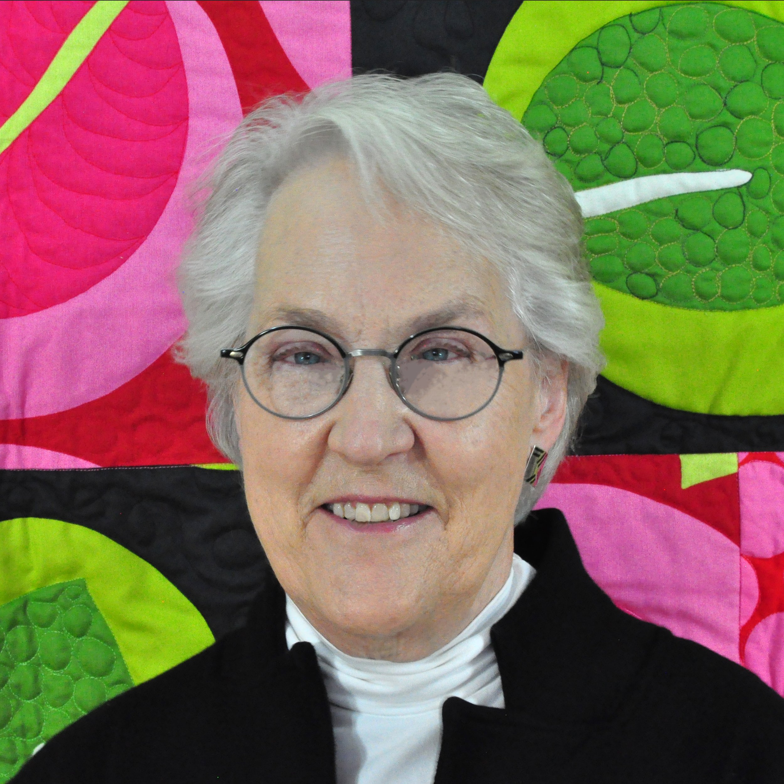 Rosalind Daniels stands in front of a multicolor abstract quilt. It has shades of bright green and pink with black in interesting patterns. Rosalind smiles, wearing glasses and a black top with white turtleneck.